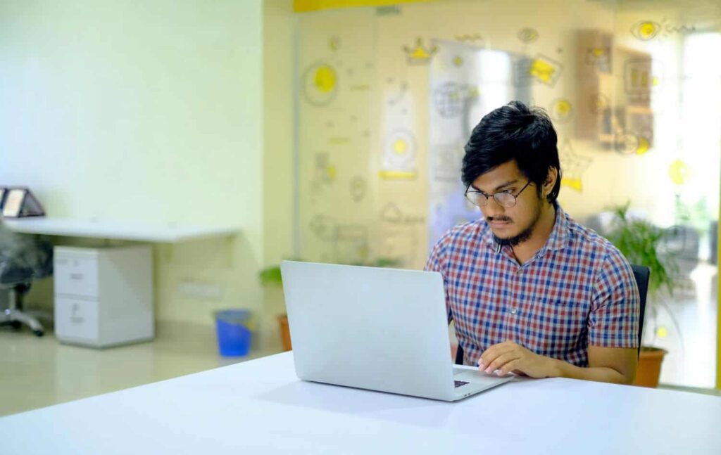 A professional sits in a bright office working on a laptop. He has glasses and dark hair and wears a checkered button-up shirt.