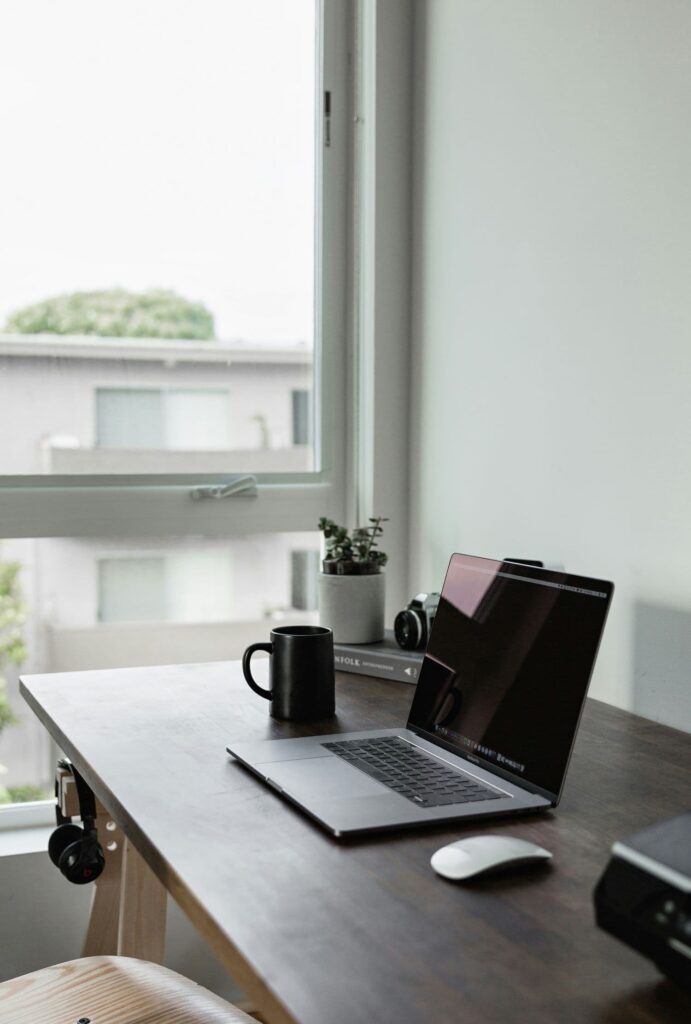 A desk with a plant, camera, coffee mug, laptop, and mouse on it. To the left is a large window overlooking other buildings.