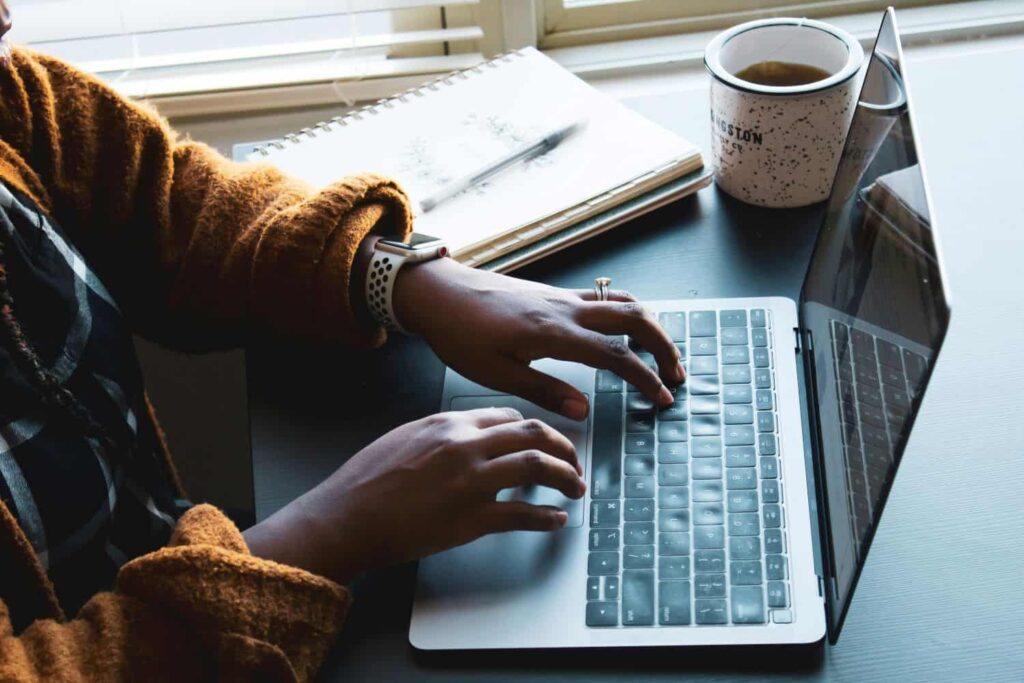 A graduate applicant sits at a desk and types an application essay on their laptop. They wear an orange cardigan and an engagement ring and have a notebook and coffee mug to their left.