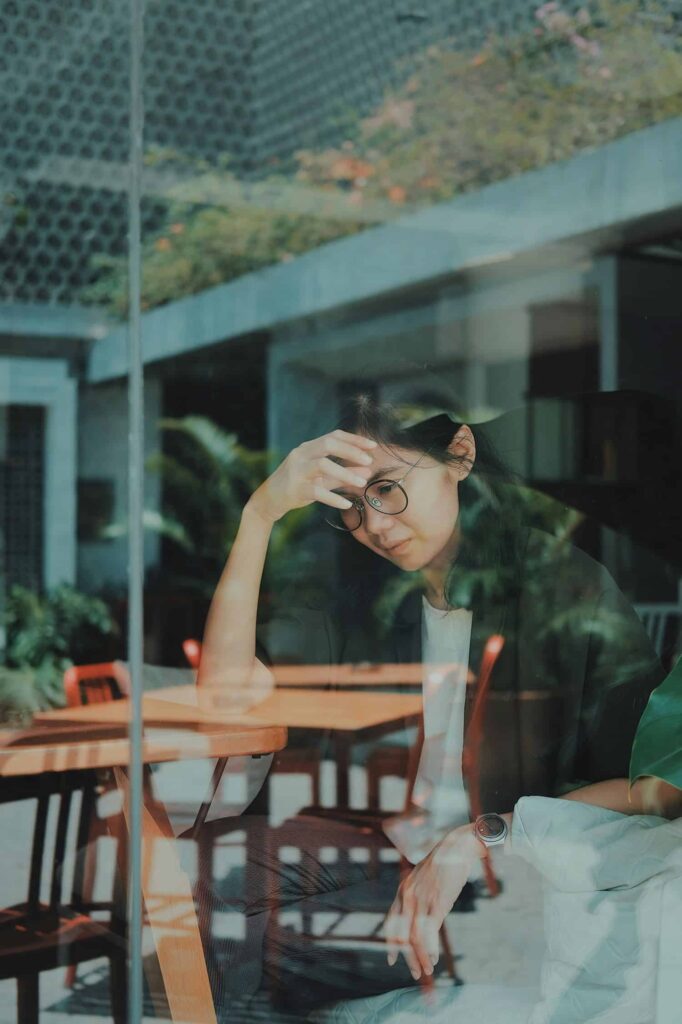 A woman looking stressed with her hand to her forehead. She is sitting in a coffee shop and wears glasses and has long dark hair