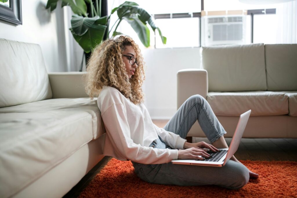 A graduate student sits on the floor of their living room working on a laptop. They wear jeans and a white pull-over hoodie and have curly, long blonde hair.