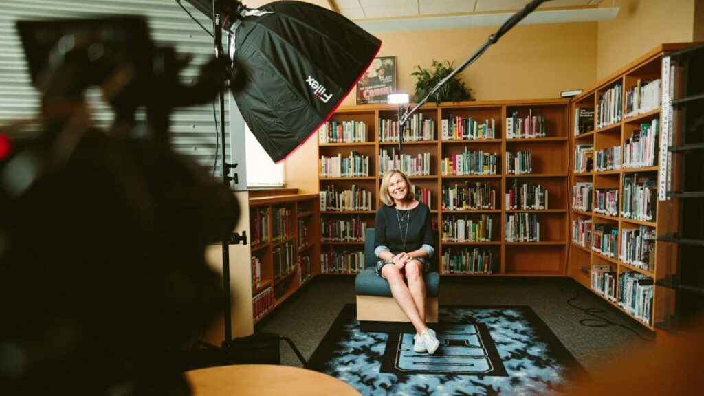 A person being interviewed in a small library. They wear a black dress and have blonde shoulder-length hair.