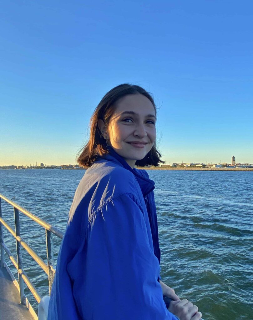 Monica Apreutesei stands on a pier at the ocean. She wears a blue jacket and has short brown hair. She smiles at the camera