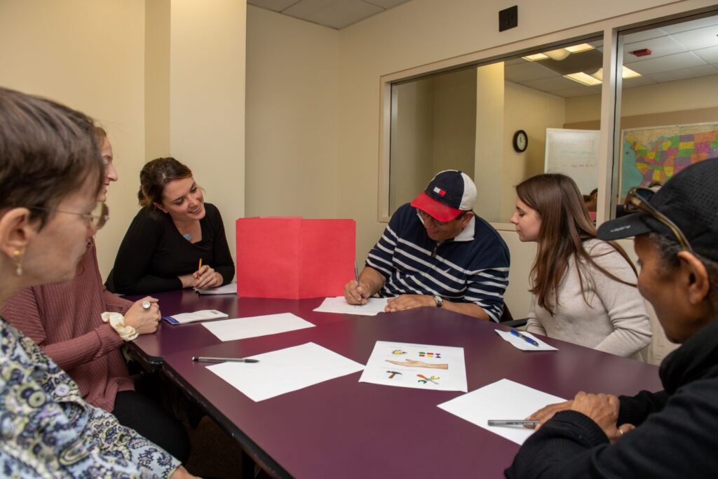 Three graduate students and three clients sit at a large table completing an activity together. At the back wall is a two-way mirror, through which the students' supervisor observes the session