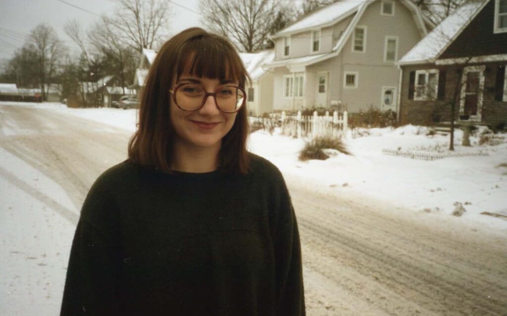 Olivia Wachtel stands by a snowy street with houses and trees in the background. She wears a dark sweater and smiles at the camera