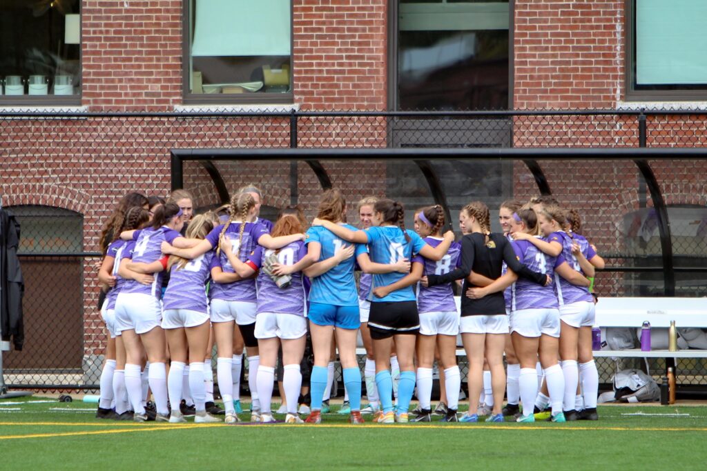 The Emerson women's soccer team huddles up on the field in an embrace.