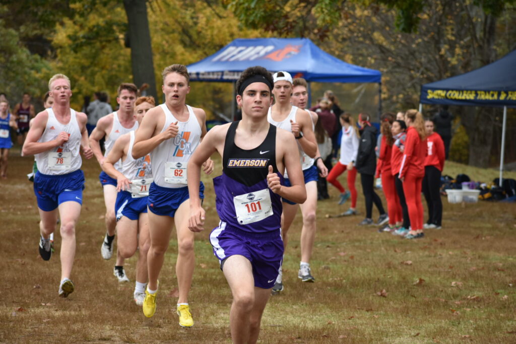 A male Emerson cross country runner runs in front of the pack at a race.