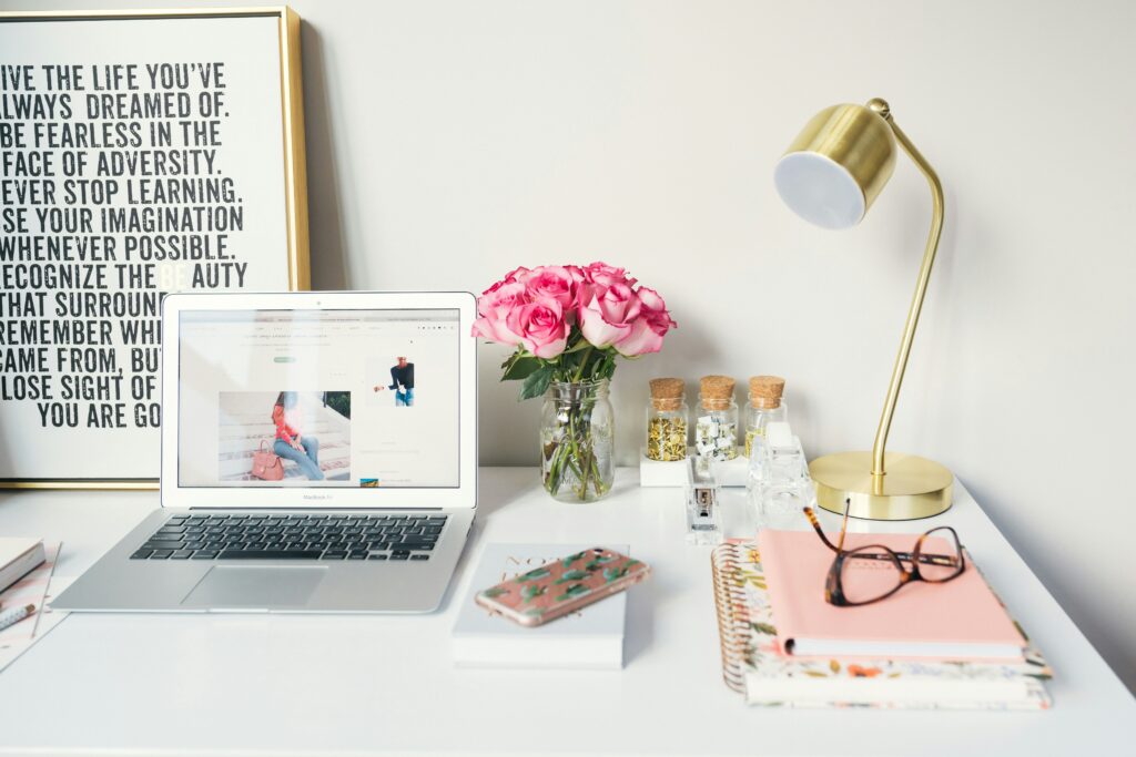 Open computer on a white desk next to flowers, notebooks with a pair of classes, and a desk lamp.