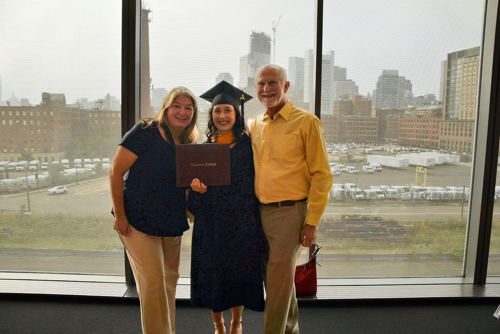 Family poses in front of large window that looks out to Boston as the graduate holds out her diploma.