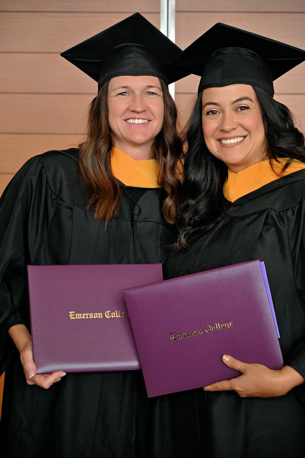 Two graduates smile at the camera with their diplomas.