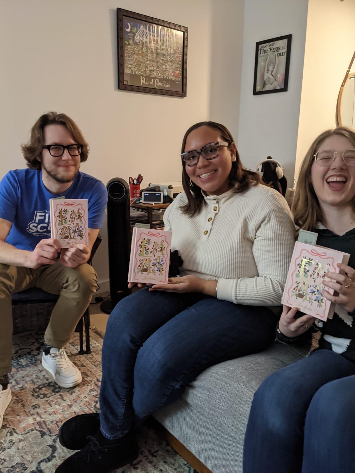 Three students smile enthusiastically while holding up their current month's book club book.