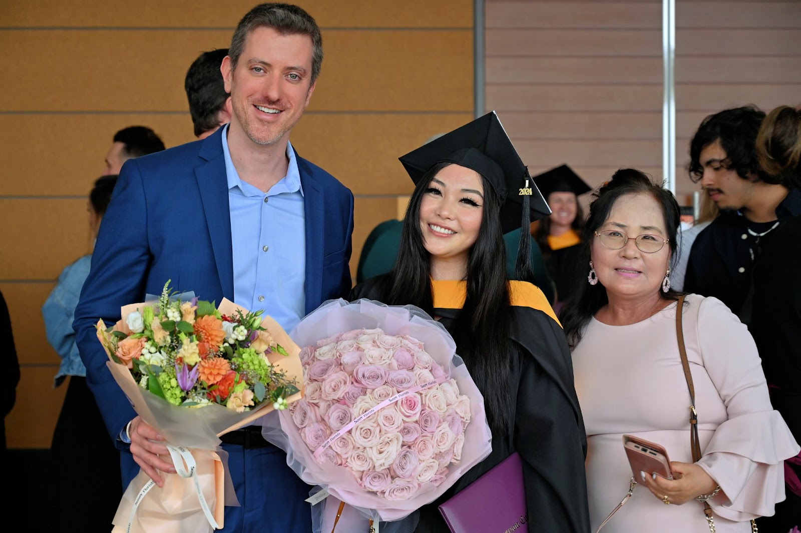 Graduate holds two bouquets of flowers as she smiles at the camera beside two of her supporters.