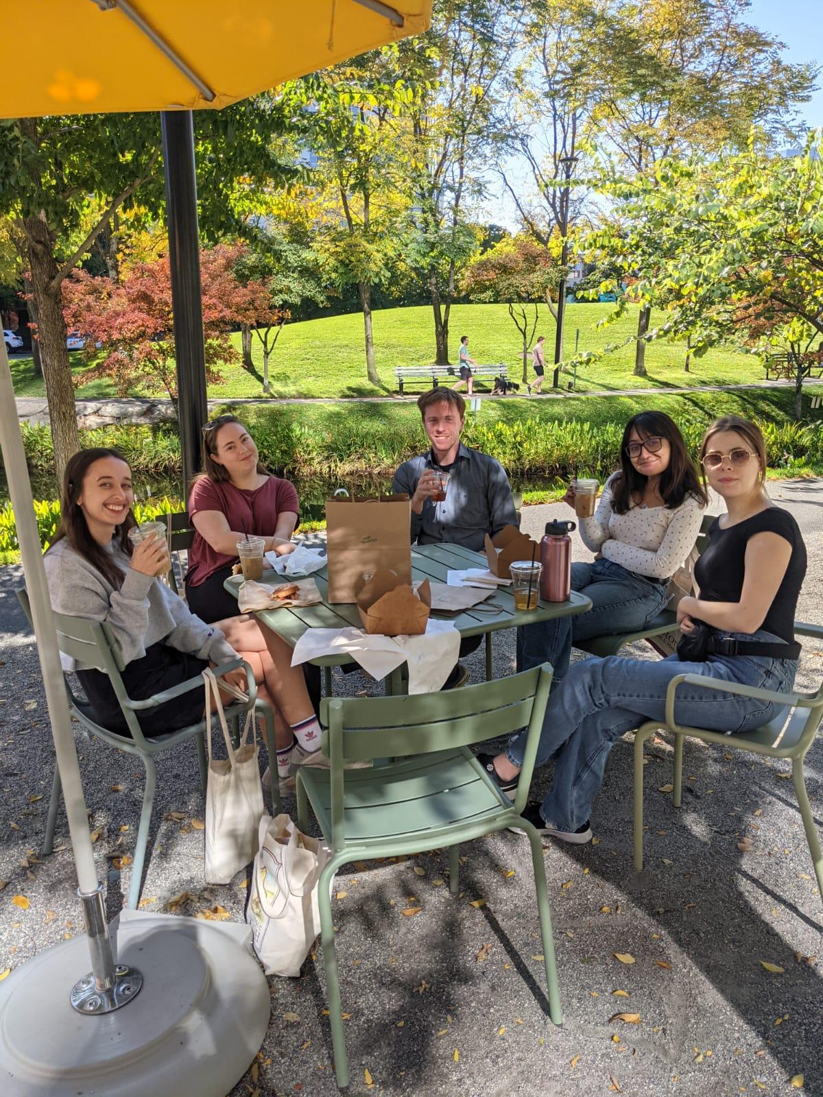 Five students sit outside at a table eating food on a warm summer day.