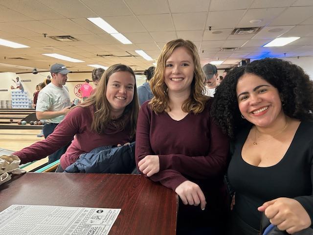 Three female students smile at the camera inside the bowling alley.