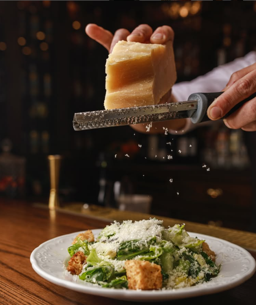 Waiter grating parmesan cheese on top of caesar salad.