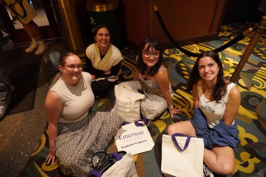 Four students sit on the floor in a theater by a sign for Emerson College graduate programs.