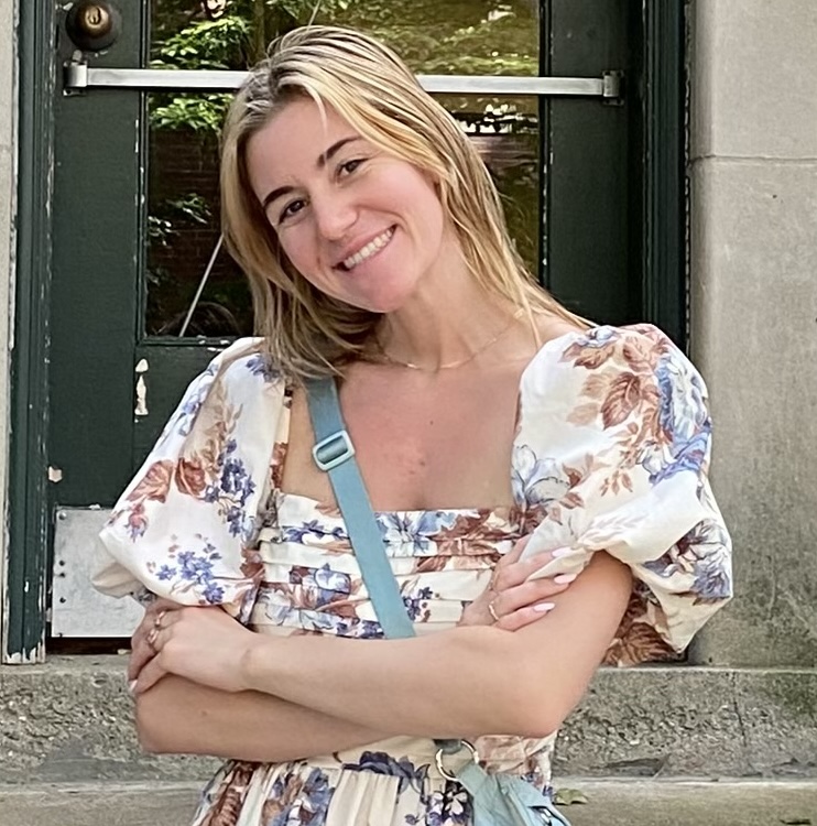 A white woman with blonde hair, Emily, smiles at the camera with her arms crossed in front of her chest in front of a green apartment building door.