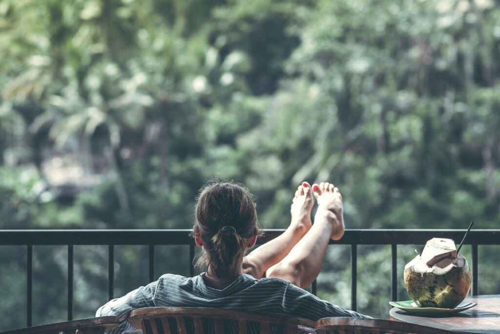 A white woman puts her feet up on a black balcony as she leans back. She has a coconut with a straw coming out of it to the right of her.