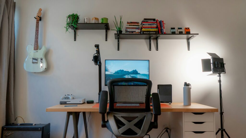 An at-home office that features a wood desk, black office chair, guitar on the wall, and shelves with plants and books.