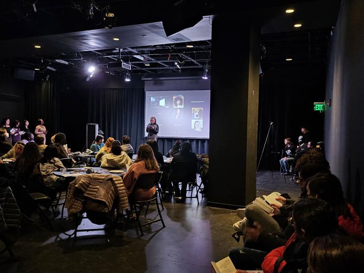 The black box theatre with a student reading their work at an open mic and viewers look on and people watch on Zoom behind the speaker.