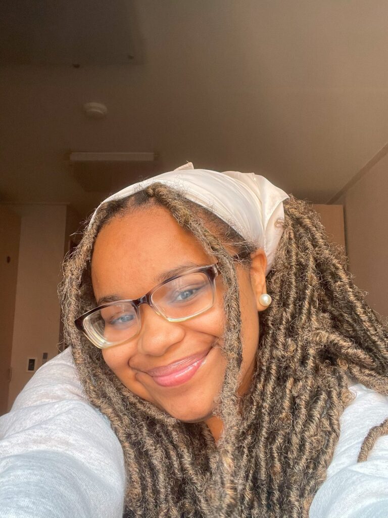 Selfie of Aniaha, a Black woman with hair in twists and a white handkerchief smiling at the camera. She has glasses and is wearing a white sweatshirt.