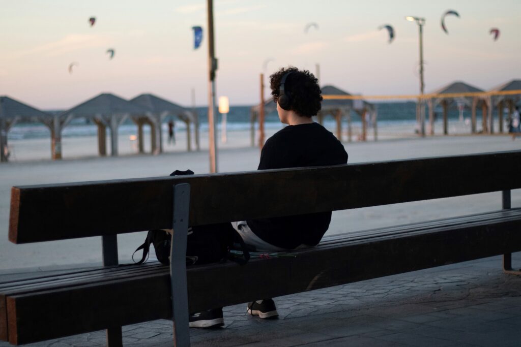 A person wearing a black sweatshirt sits on a bench and looks out. He may be listening to podcasts in his headphones.