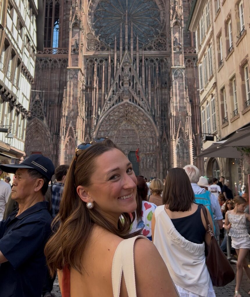 Girl smiles back at a camera in front of famous architecture. She has brown hair and is wearing sunglasses on her head with a canvas tote bag on her shoulder.