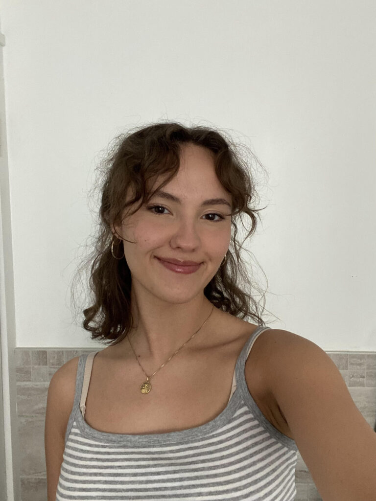 Woman smiles at camera in a selfie format. She has short, curly brown hair and is wearing a white and grey striped tank top. 