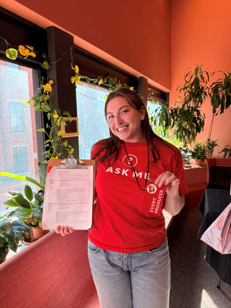 A white woman with brown hair wears a red "Ask Me" shift and poses with her "event producer" lanyard and clipboard.