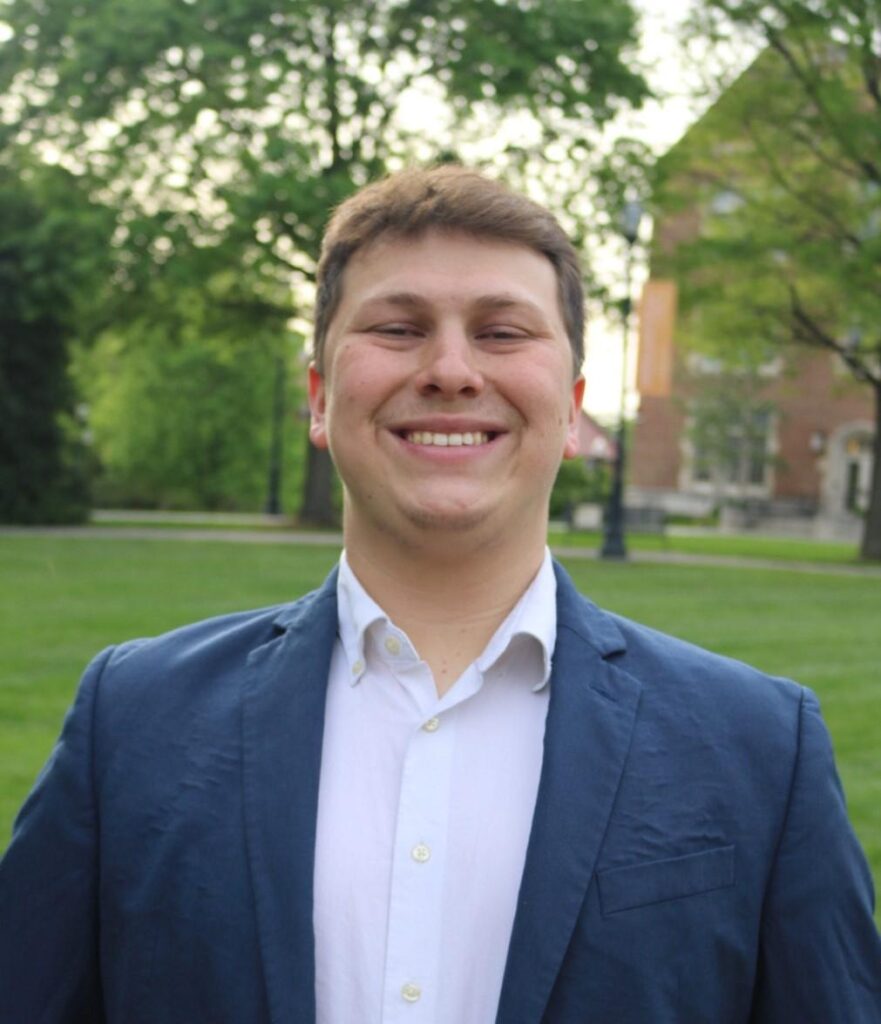 Benjamin Pinzon smiles at the camera in an outdoor portrait shot. He is wearing a navy blazer with a white collared shirt.