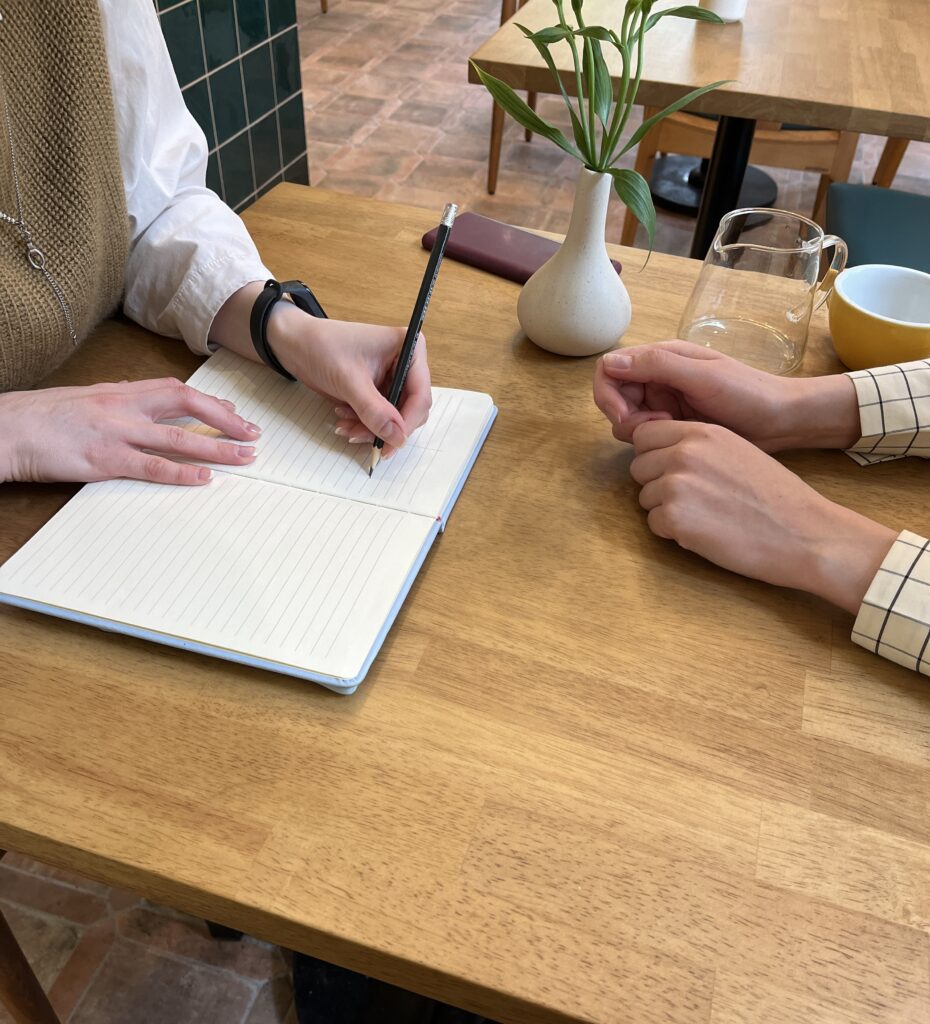 Two people sit aross from each other at a wooden table. One person is writing something with a pencil in a blue journal.