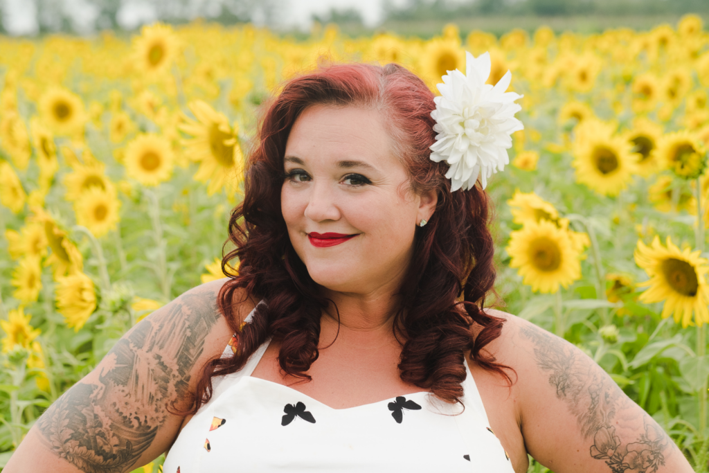 Mickie smiles at the camera as she poses with a flower in her red hair, standing in a sunflower field.