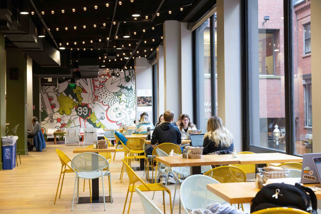 Students sit at wooden tables with yellow chairs while string lights hang from the ceiling and windows look out to the street.