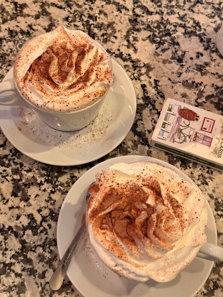 Two hot chocolates in white mugs with whipped cream and sprinkled cocoa powder sit on a table beside a deck of cards.