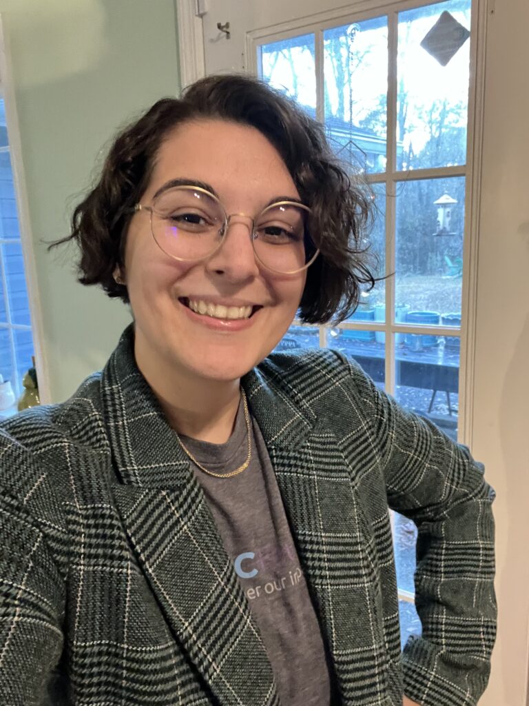 Photo of Susanne Salehi smiling at the camera. She is a white woman with short brown hair and round glasses wearing a plaid blazer.