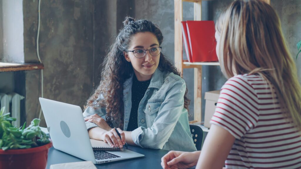 Young female entrepreneur is talking to her partner in modern office while sitting at desk together. 