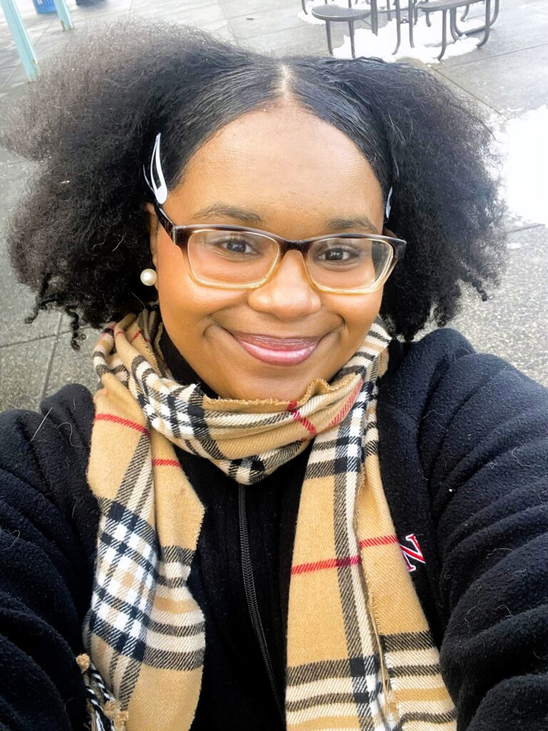 close up of woman smiling with brown skin and black curly hair. She is wearing glasses and a plaid scarf and a black sweater