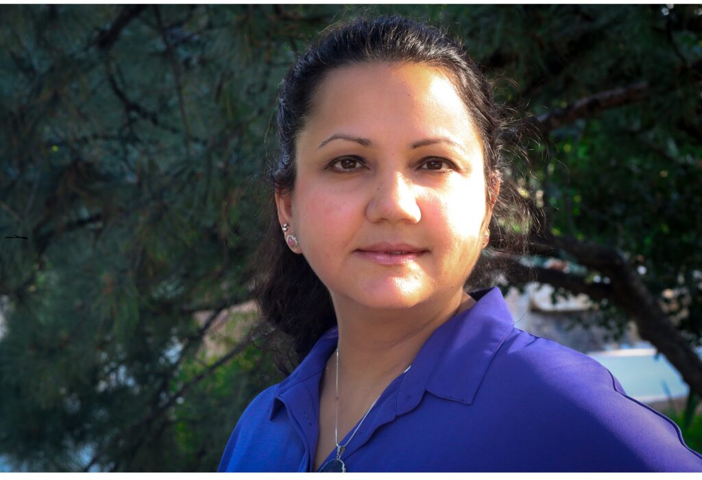close up of a woman with dark hair pulled back. She has olive skin and brown eyes. She is wearing a blue shirt