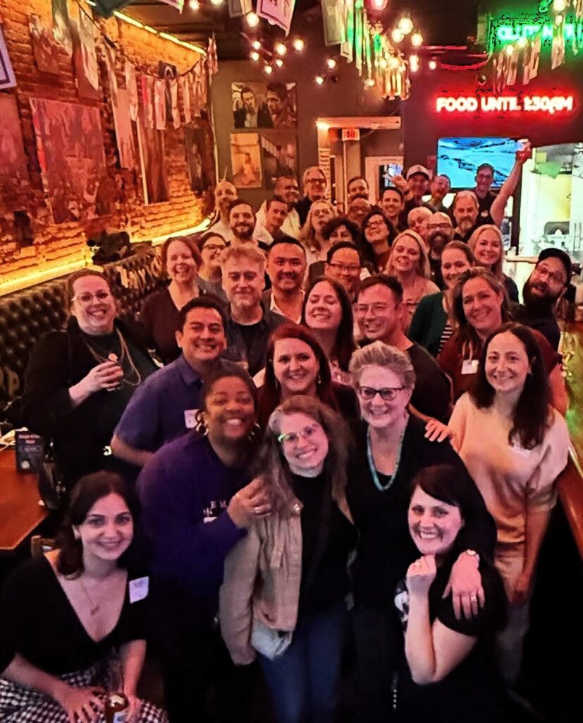 photo of group of smiling young adults in colorfully lit bar surrounding an older woman wearing black framed glasses and smiling.