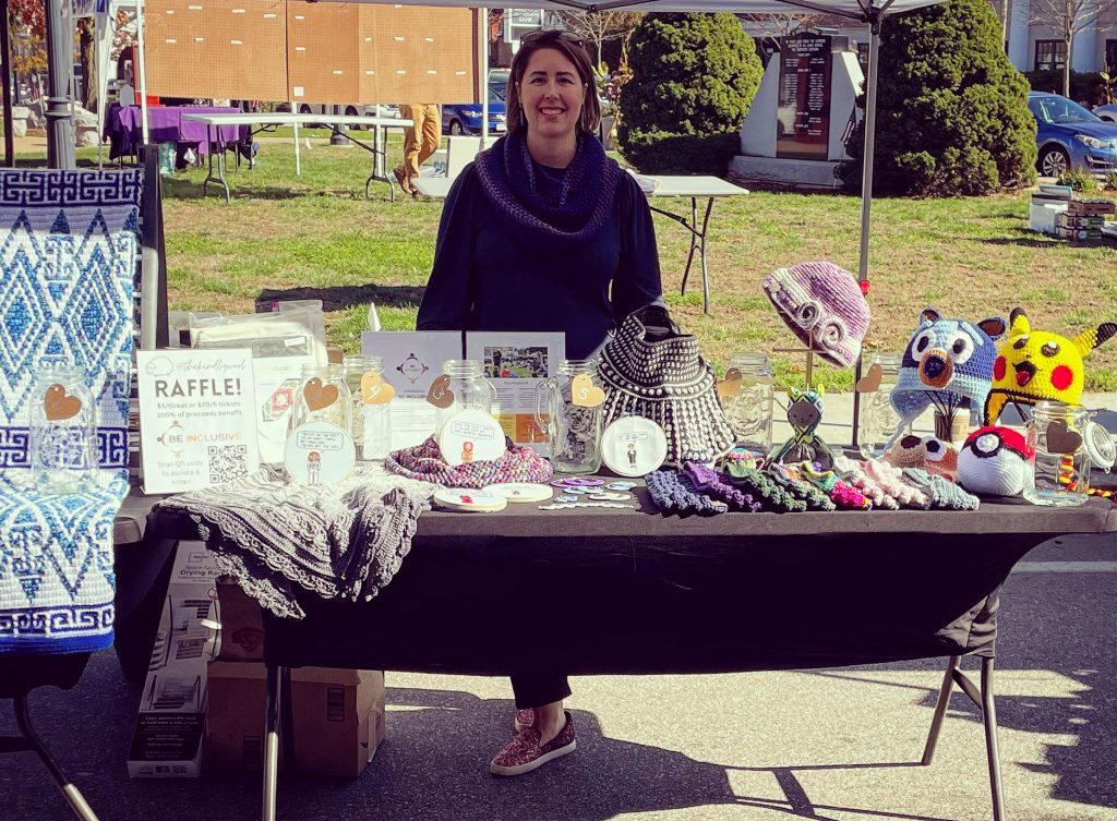 Liz Chase stands at a table with crocheted items to raffle to benefit Be Inclusive