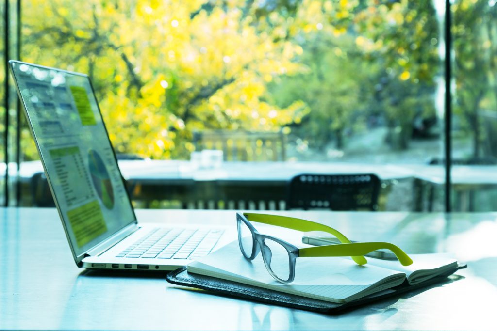 Laptop, notebook and eyeglasses rest on a table in front of a large window