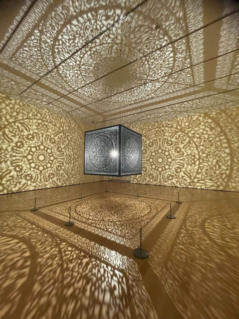 It’s a light-based exhibit where a bright lamp inside a decorative metal cage casts shadows on all walls, floor, and ceiling of a bare, cream room. The shadow designs are floral with organic swirls and patterns, organized in concentric circles of expanding motifs.